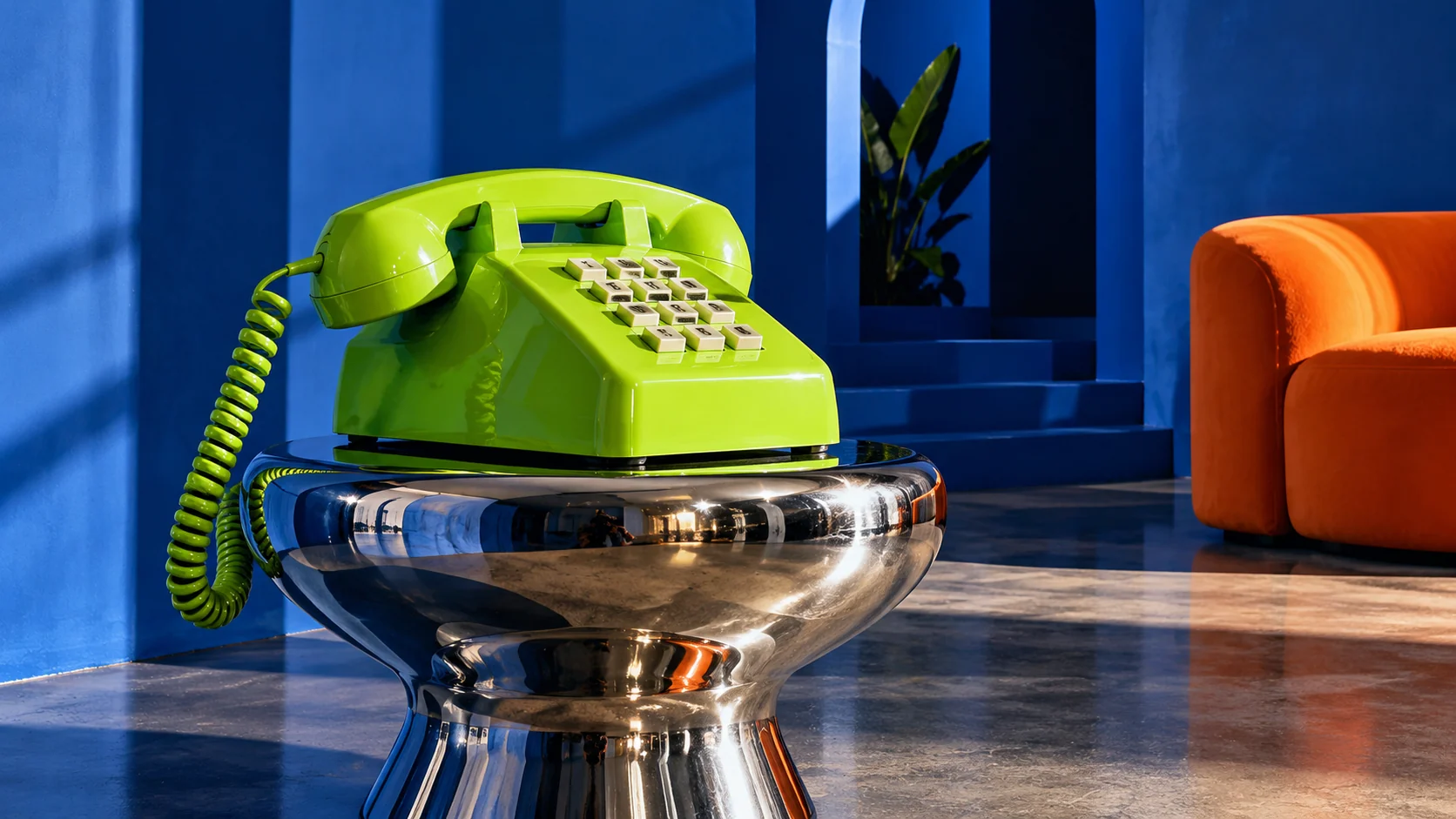 A lime-green retro telephone on a polished chrome table in a cobalt-blue room with an orange sofa